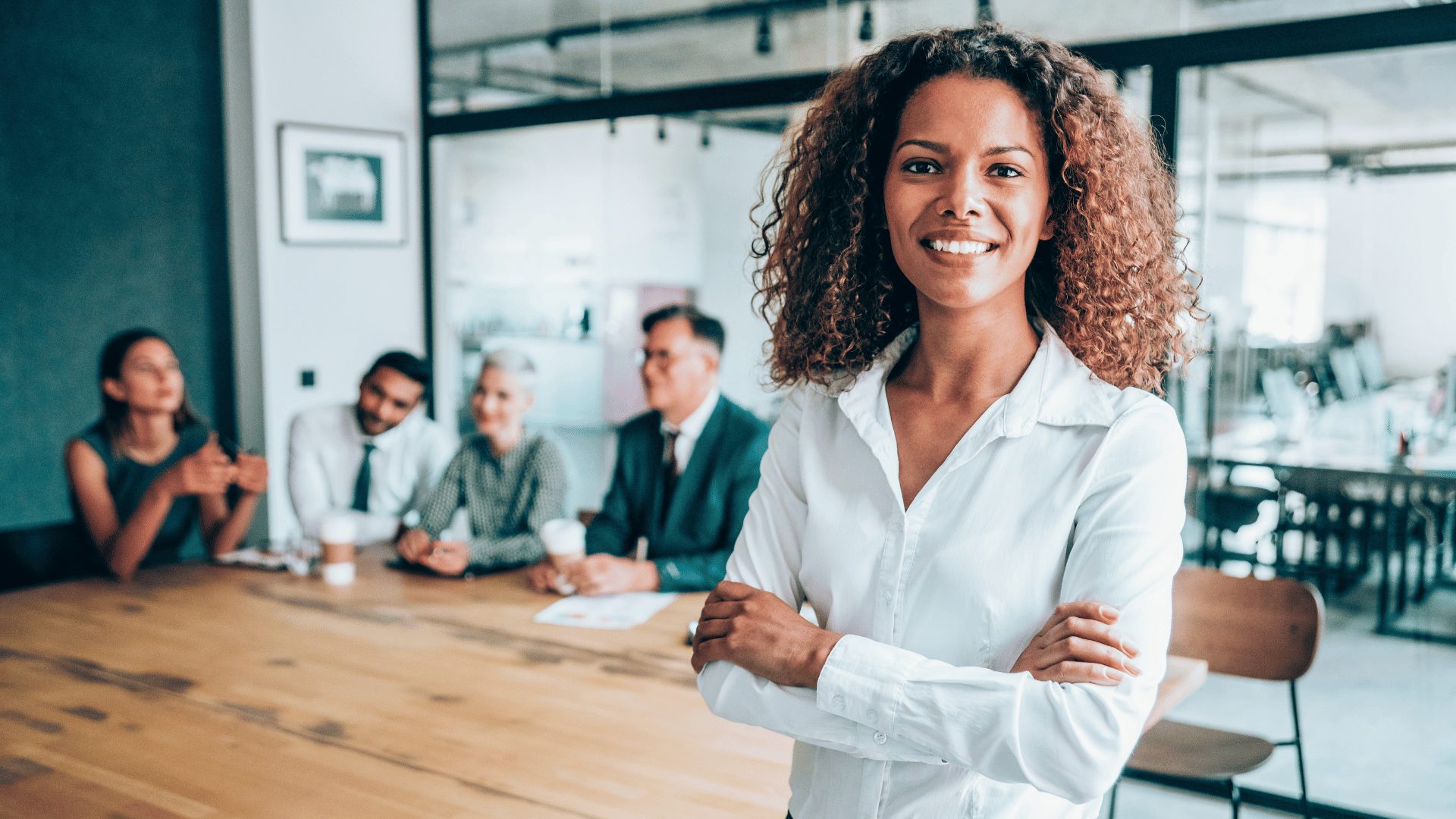 A female senior leader, smiling at the camera, stood in front of a desk with her colleagues sat behind her.