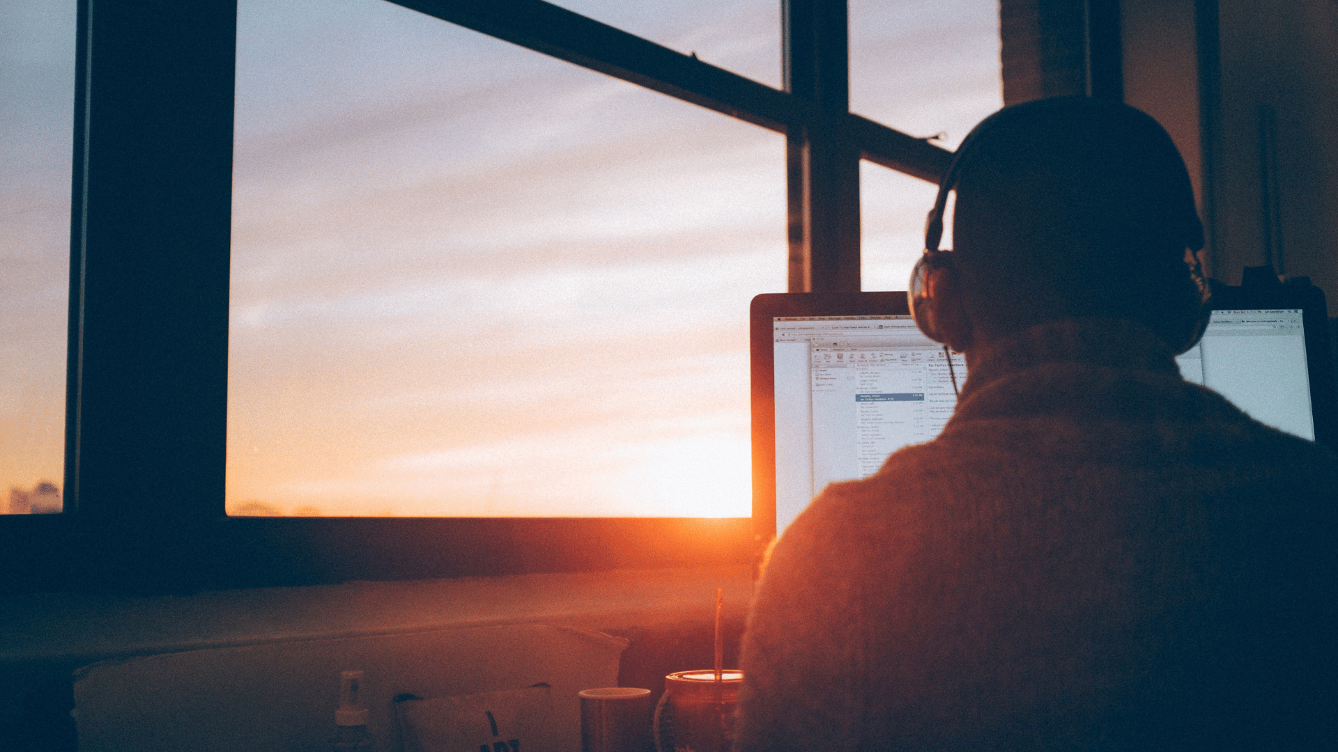 A man studying at a computer in front of a big glass window as the sun sets